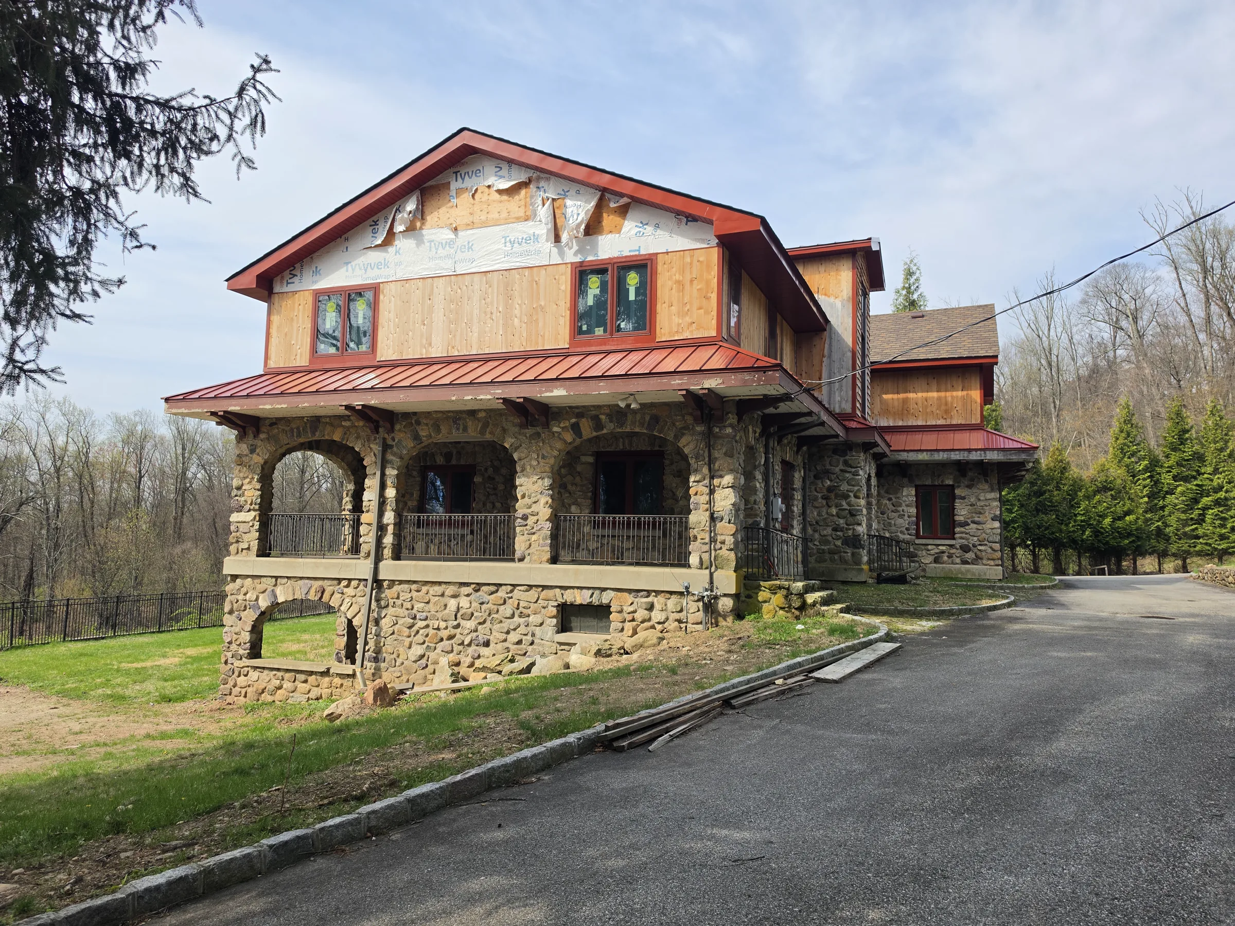 River-stone house under construction with a new cedar-clad second story, exposed house wrap at the gable peak, and a red standing-seam porch roof